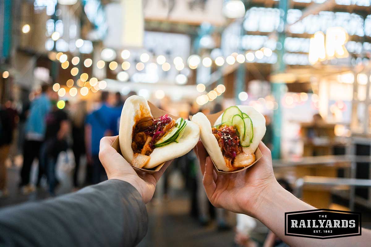 Two bao sandwiches are in the foreground; the background shows an out-of-focus food festival.