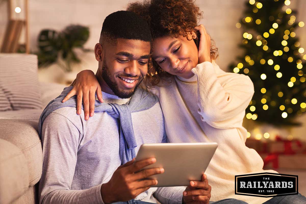 A couple sitting on their couch together in front of a Christmas tree, looking at their tablet. Discover safe Sacramento holiday events for 2020.